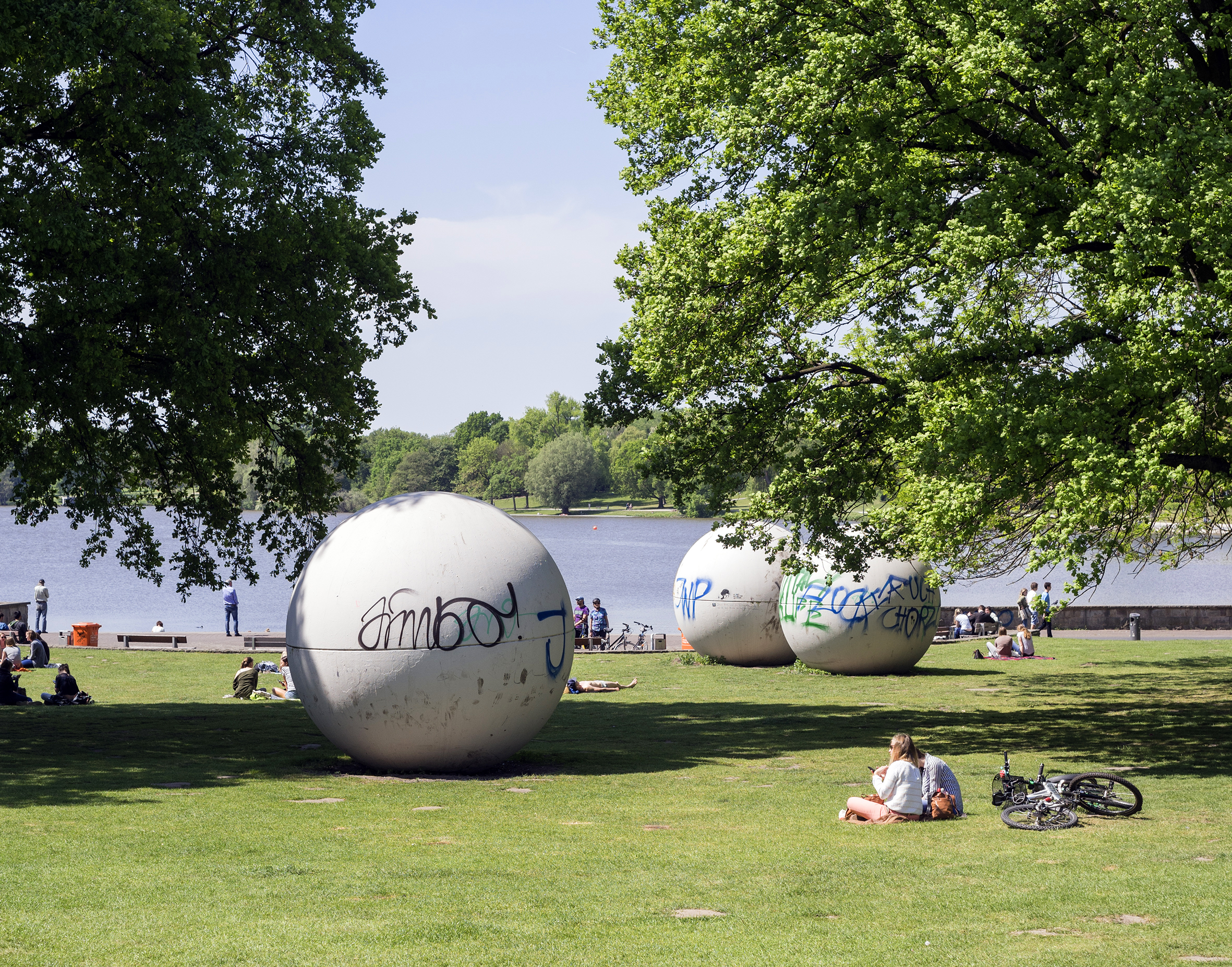 Das Foto zeigt große, weiße Betonkugeln auf einer Wiese. Im Hintergrund ist ein See zu sehen und im Vordergrund zwei große, grüne Bäume.