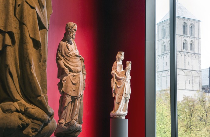 Das Foto zeigt mittelalterliche Skulpturen in einem rot gestrichenen Raum. Aus dem Fenster heraus sieht man den Münsteraner Dom.