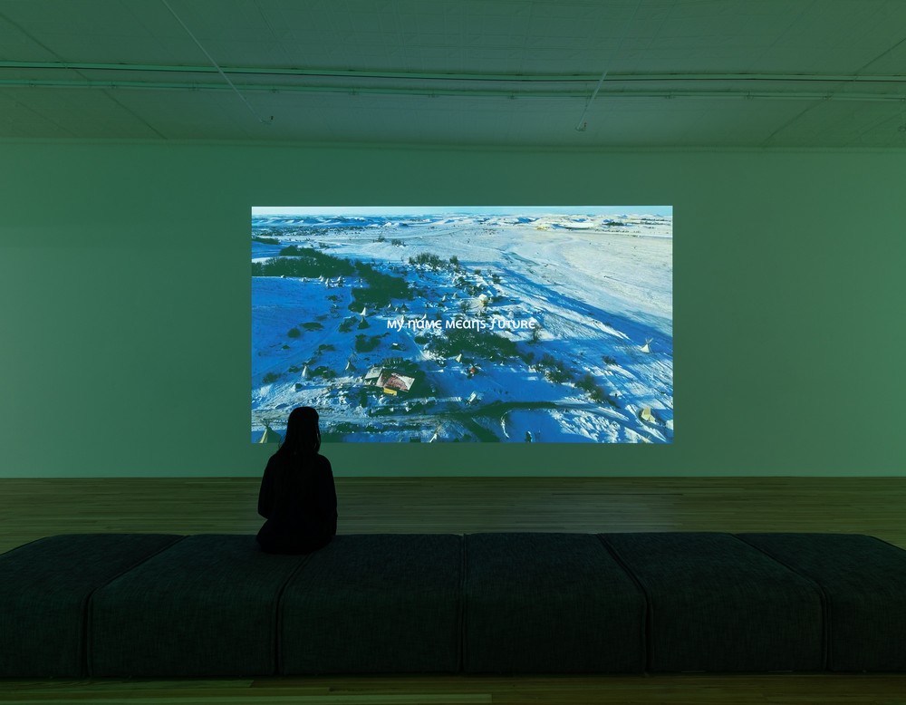Eine Frau schaut auf einem Bildschirm an einer Wand. Das Bild zeigt eine Schneelandschaft und den Titel "My name means future"