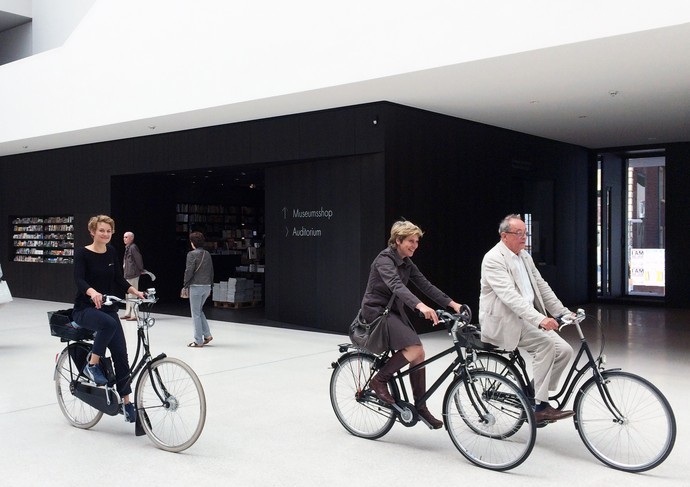 Kasper König, Britta Peters und Marianne Wagner fahren mit dem Fahrrad durch das Foyer des Museums.