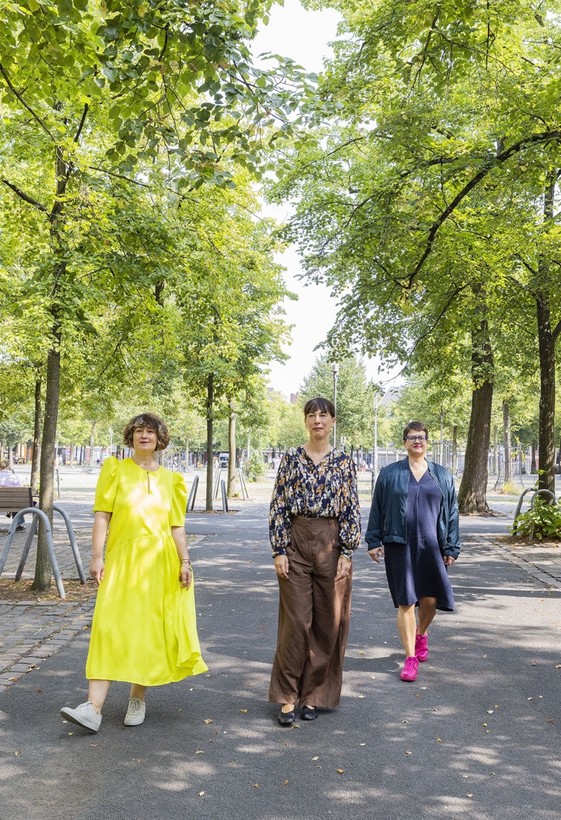 Three women are standing on a street. You can see green trees in the background.