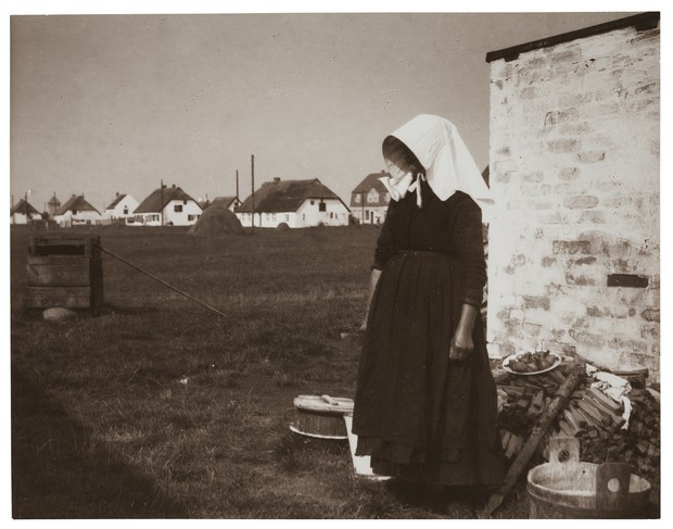 Frau mit weißer Haube vor einer Steinmauer, im Hintergrund Wohnhäuser.