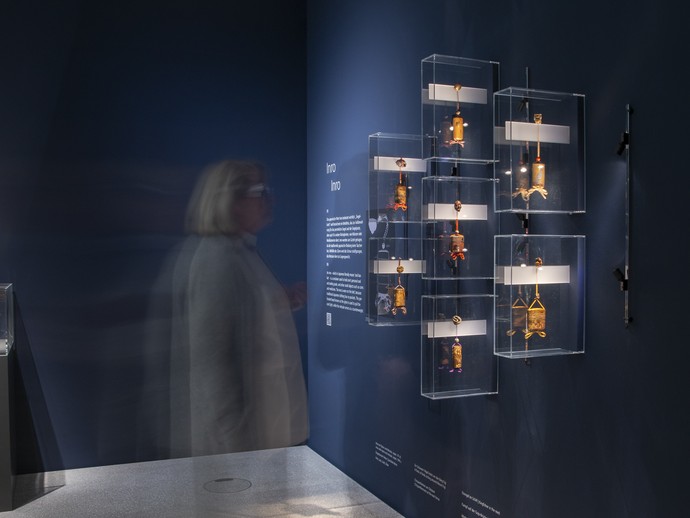 Das Bild zeigt eine Aufnahme aus der Ausstellung. Zu sehen ist ein blauer Raum mit einer Vitrine, in der sich kleine Objekte befinden. (vergrößerte Bildansicht wird geöffnet)