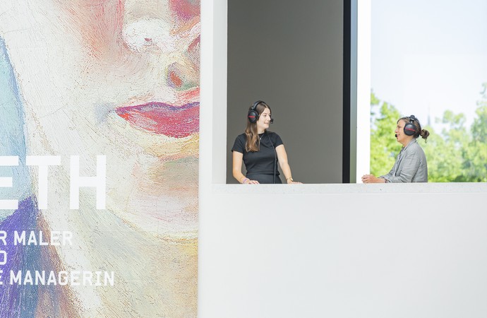 Two women stand with headphones next to the exhibition poster of Elisabeth Macke.