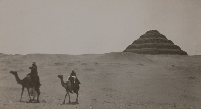 Zwei Kamelreiter in der Wüste, im Hintergrund Pyramiden.