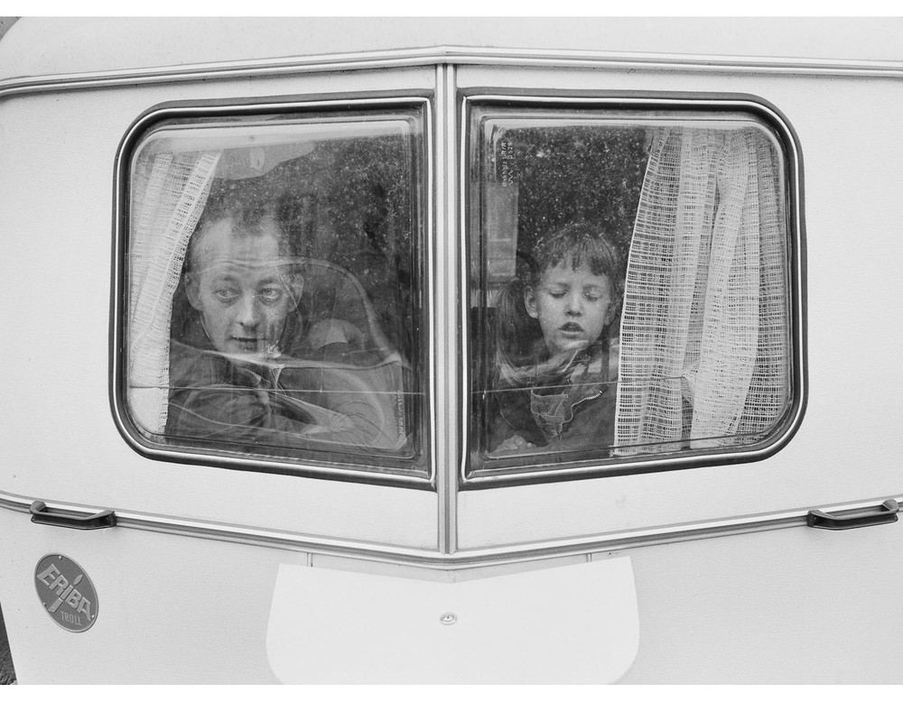 Das schwarz-weiß Foto zeigt einen Mann und ein Kind, die aus dem Fenster eines Wohnwagens schauen.