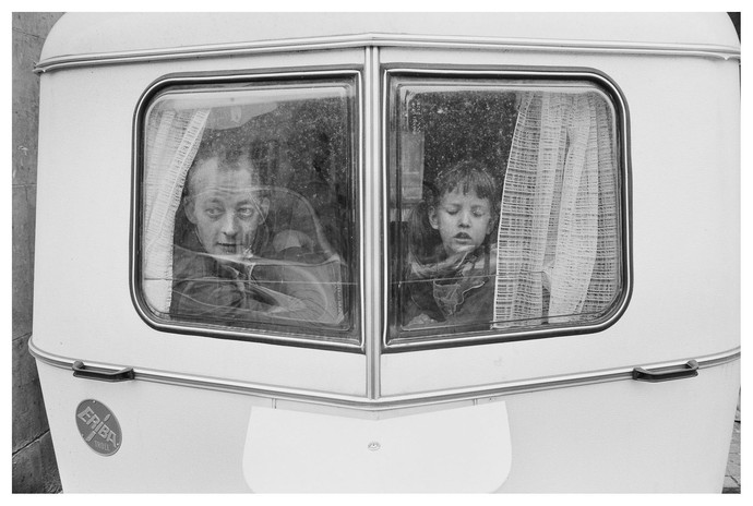 Das schwarz-weiß Foto zeigt zwei Personen, ein Mann und ein Kind, die aus dem Fenster eines Wohnwagens schauen.
